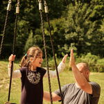 Ein lachendes Maedchen auf einer Schaukel gibt einem Erwachsenen freudig ein High-Five, waehrend eine weitere Person daneben steht - eine Szene voller gemeinsamer Freude und Teilhabe in der Natur.